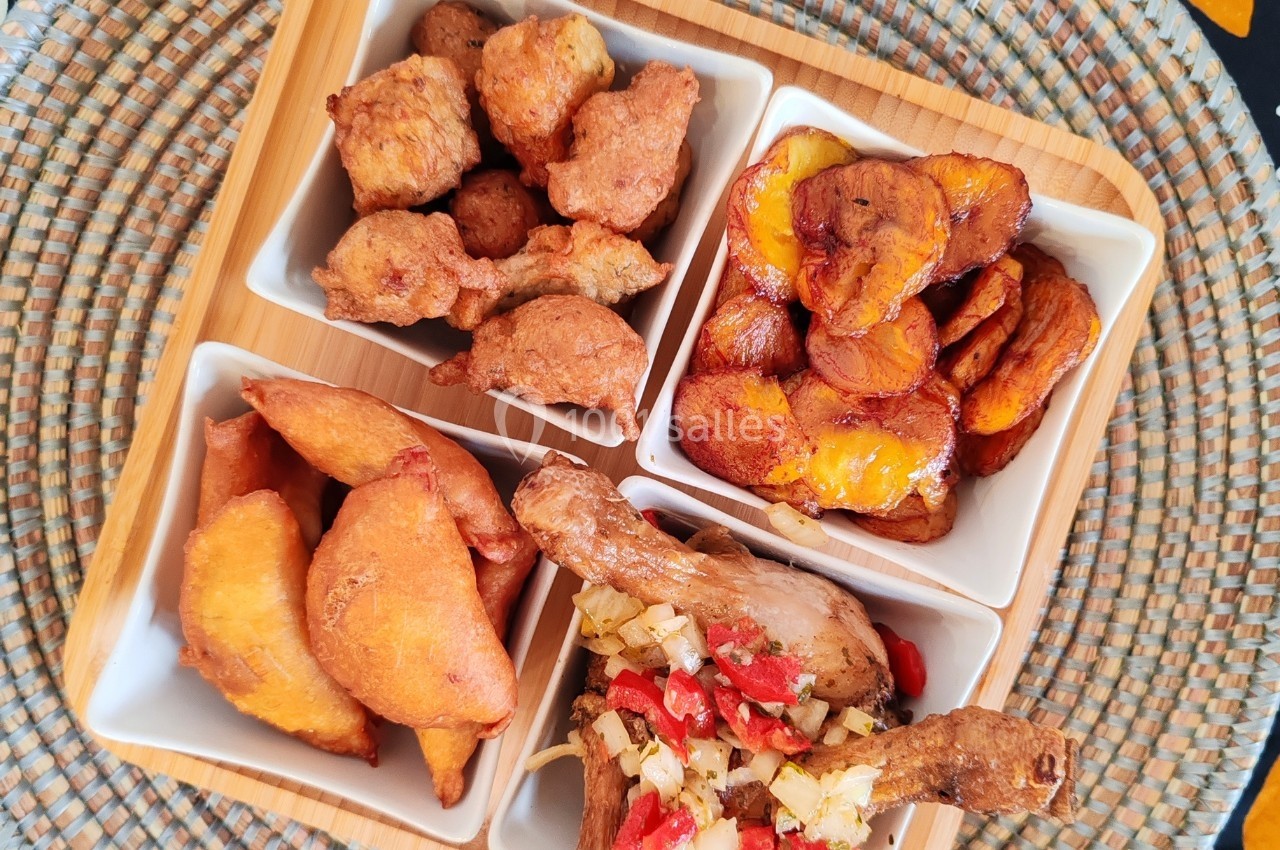 Assortiment de beignets, bananes plantains frites et poulet épicé servi dans des bols sur un plateau en bois.