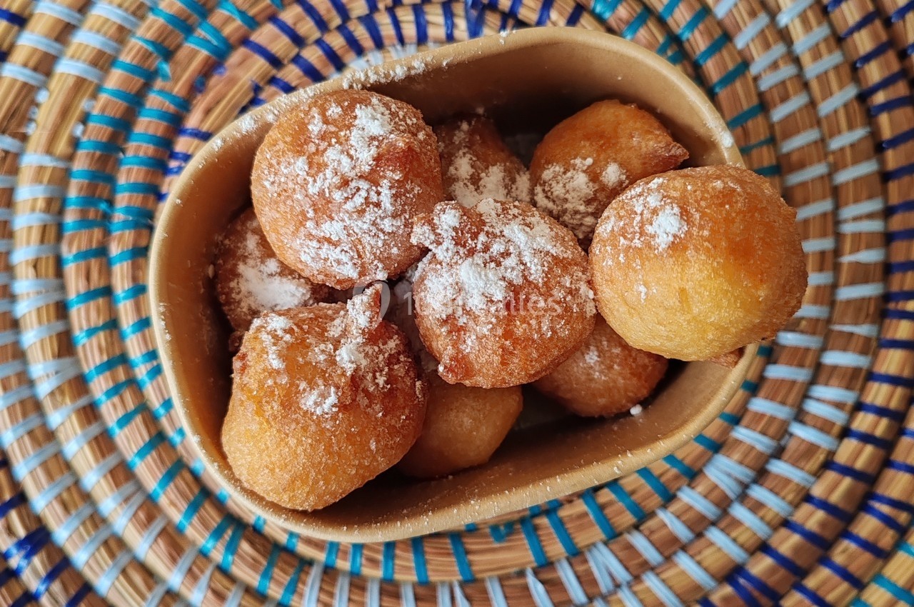 Beignets dorés saupoudrés de sucre glace dans un bol, posés sur un plateau en osier coloré.
