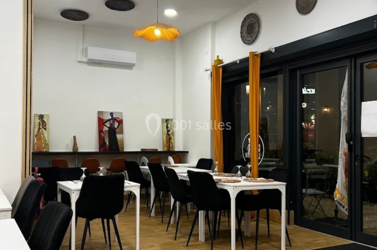 Salle de restaurant moderne avec chaises noires, tables blanches, décorations murales et grandes baies vitrées.