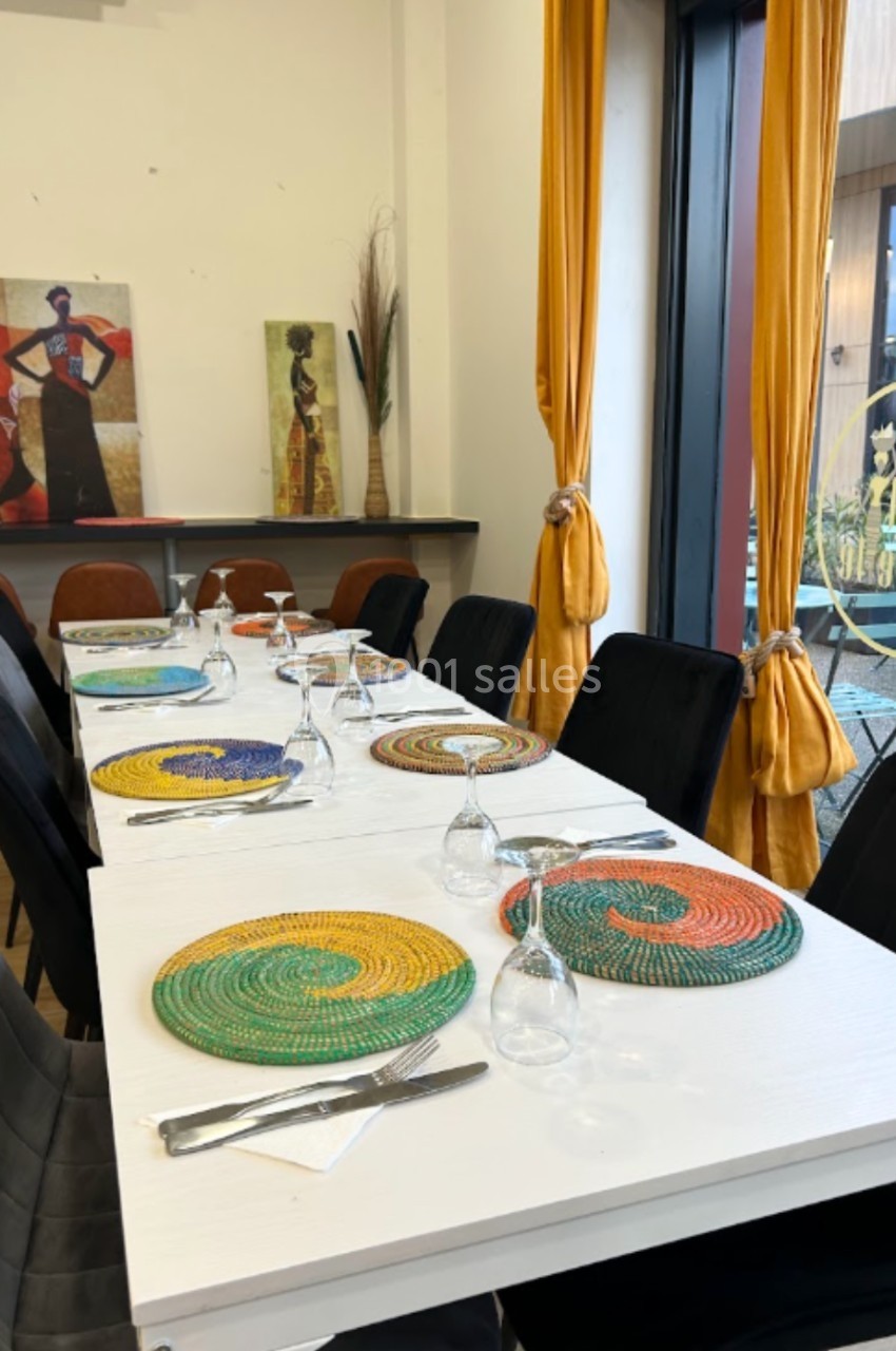 Table dressée avec des assiettes colorées tressées, verres et couverts, dans une salle lumineuse avec rideaux jaunes.