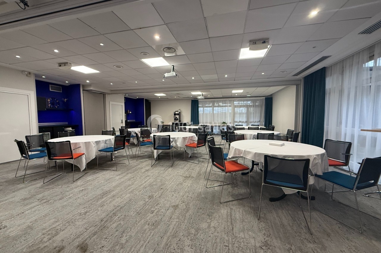Salle de réunion lumineuse avec tables rondes dressées de nappes blanches et chaises colorées.