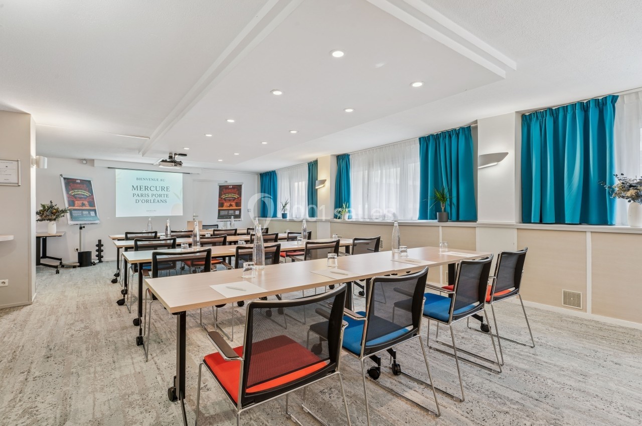 Salle de réunion lumineuse avec tables, chaises, écran de projection et rideaux bleus.