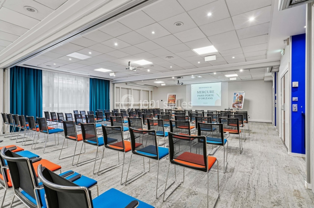 Salle de conférence lumineuse avec des rangées de chaises colorées, écran de projection et rideaux bleus.