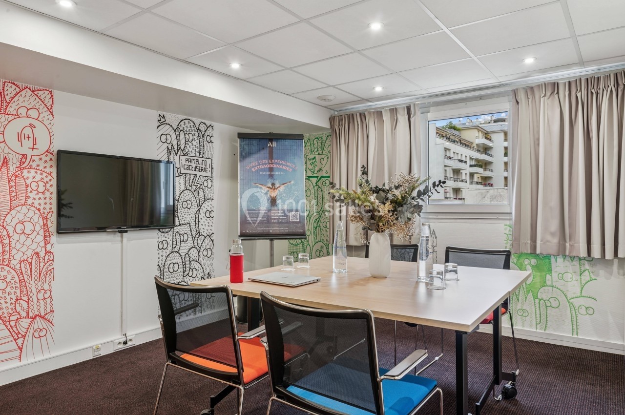 Salle de réunion lumineuse avec table, chaises, écran mural, affiches décoratives et fenêtre donnant sur des immeubles.