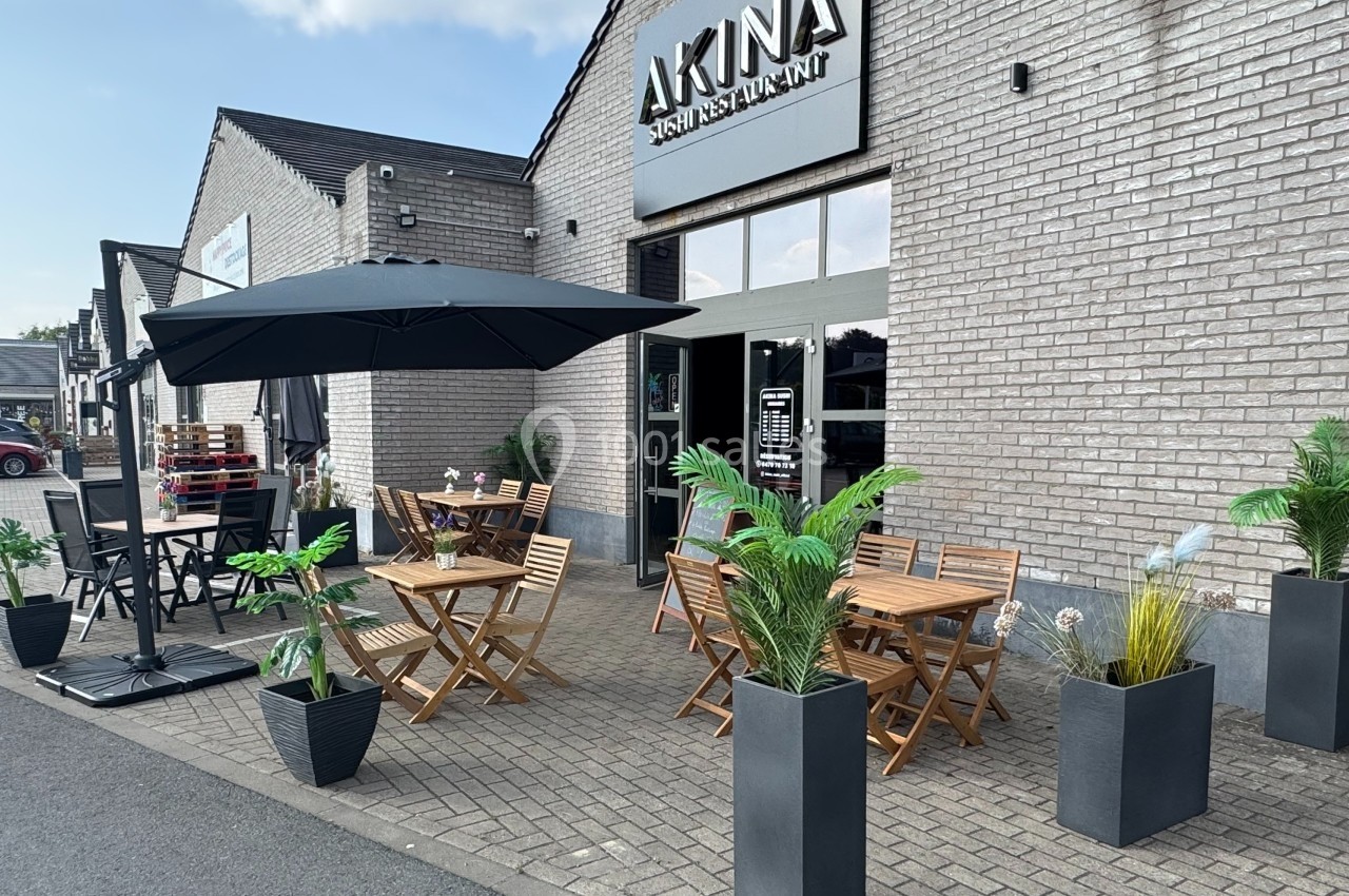 Terrasse d'un restaurant avec tables en bois, parasols noirs et plantes en pots devant une façade en briques grises.