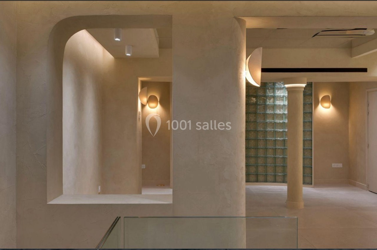 Intérieur minimaliste avec murs beige texturés, colonnes, éclairage mural et cloison en briques de verre.