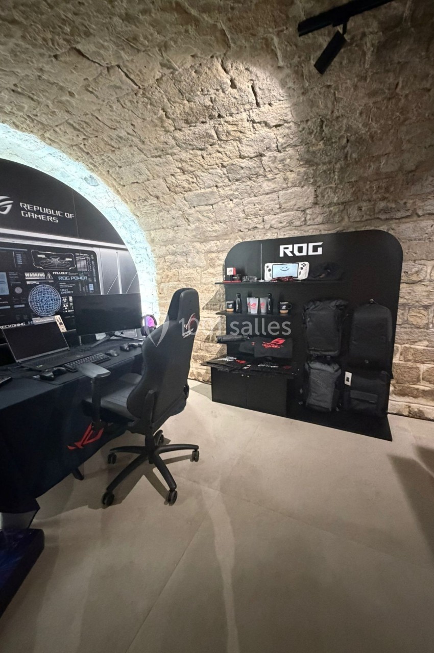 Poste de travail gaming avec chaise, écran et accessoires, situé dans une pièce voûtée en pierre avec présentoir de sacs et…