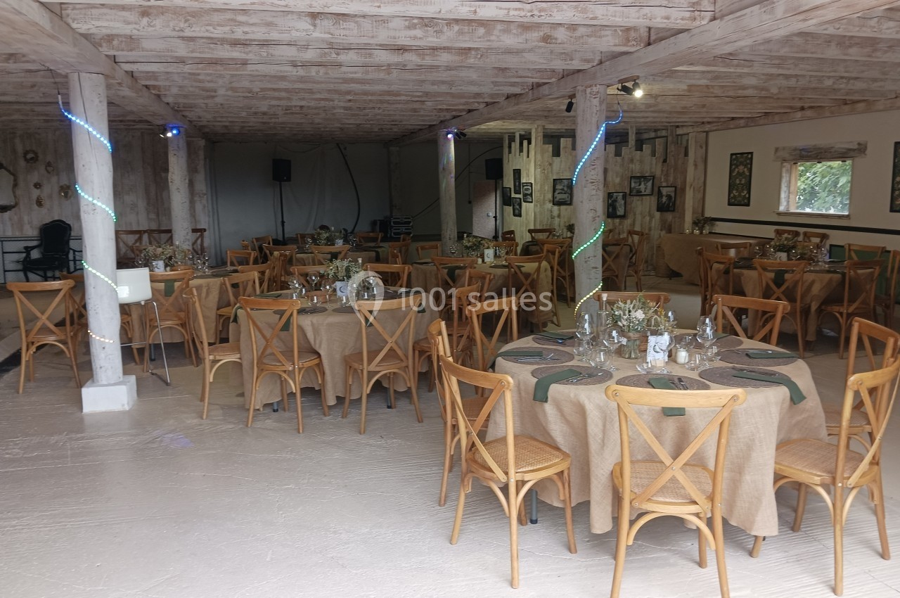 Salle de réception avec tables rondes dressées, nappes en toile de jute et chaises en bois, éclairage discret.