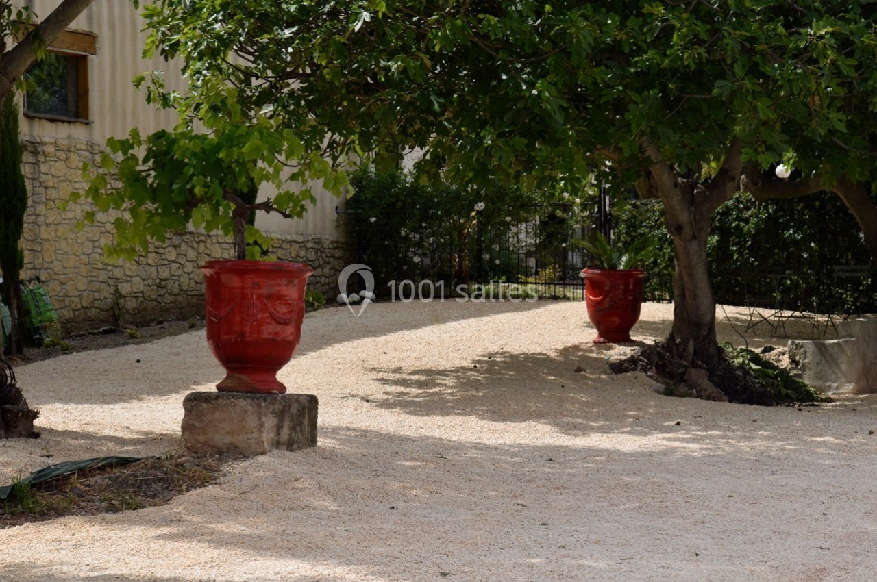 Deux grands pots rouges décoratifs placés sous des arbres dans une cour en gravier, près d'un mur en pierre.