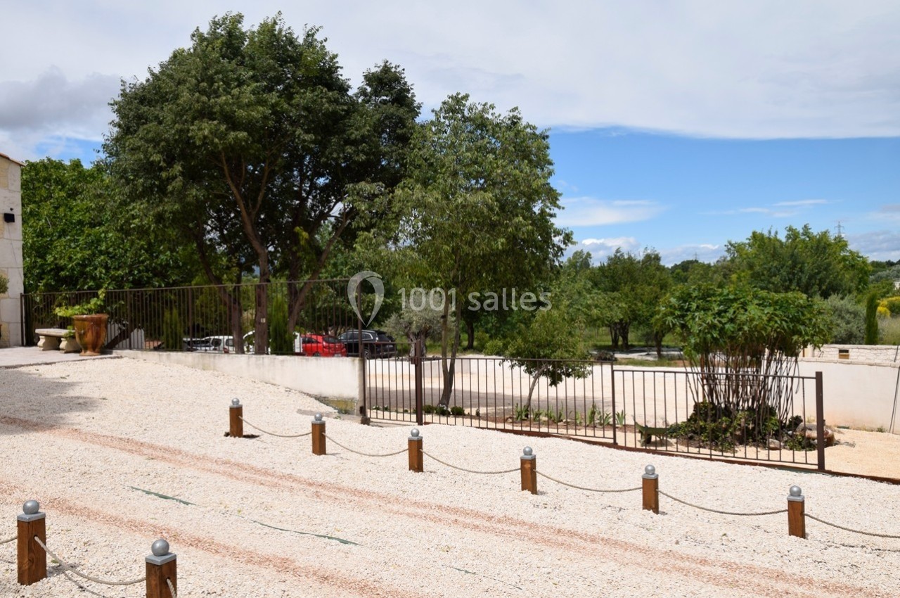 Cour extérieure avec sol en gravier, clôture métallique, arbres et quelques voitures garées en arrière-plan.