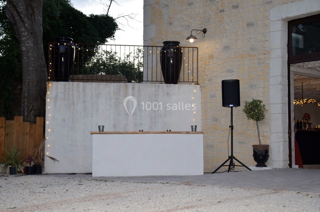 Terrasse en pierre avec un comptoir blanc, des guirlandes lumineuses, des pots noirs et un haut-parleur à droite.