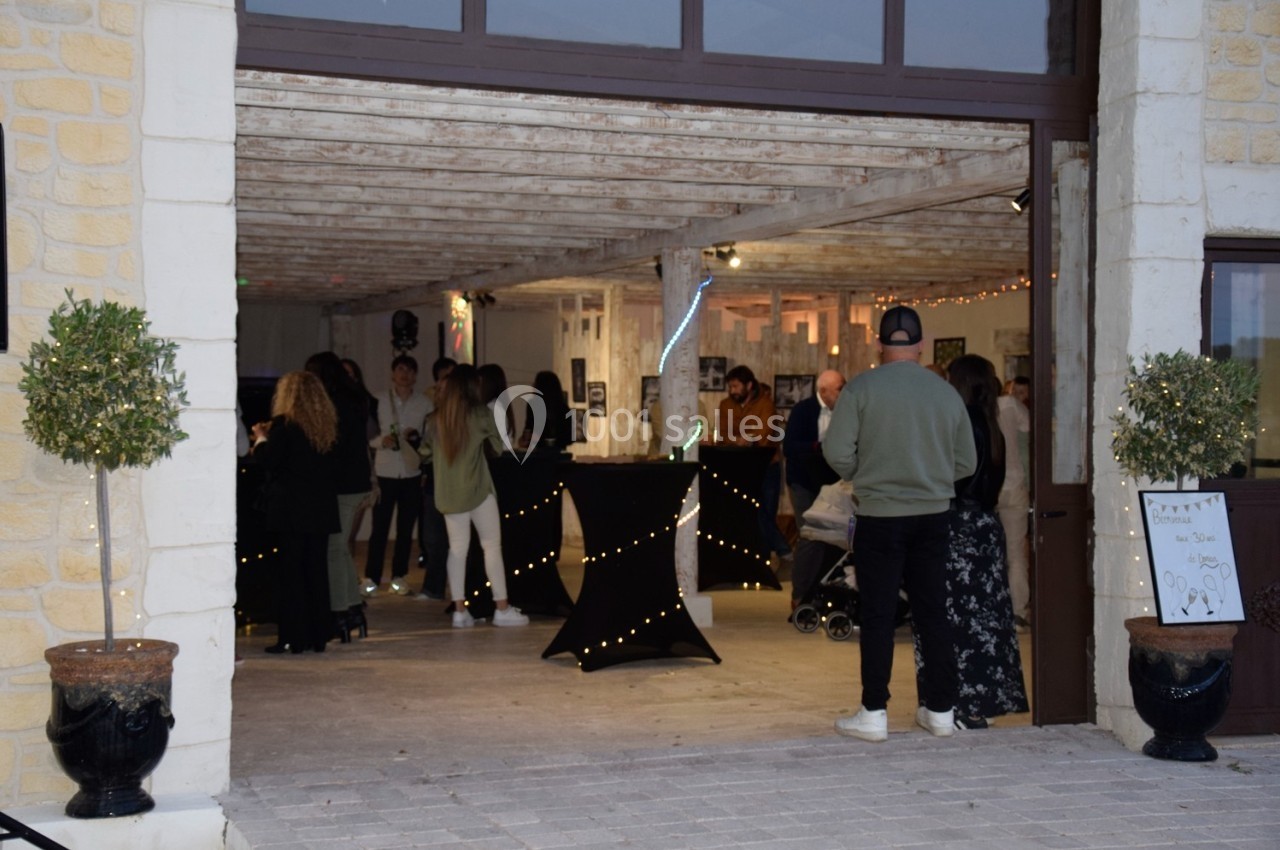 Entrée d'une salle de réception avec des invités debout près de tables décorées de guirlandes lumineuses.