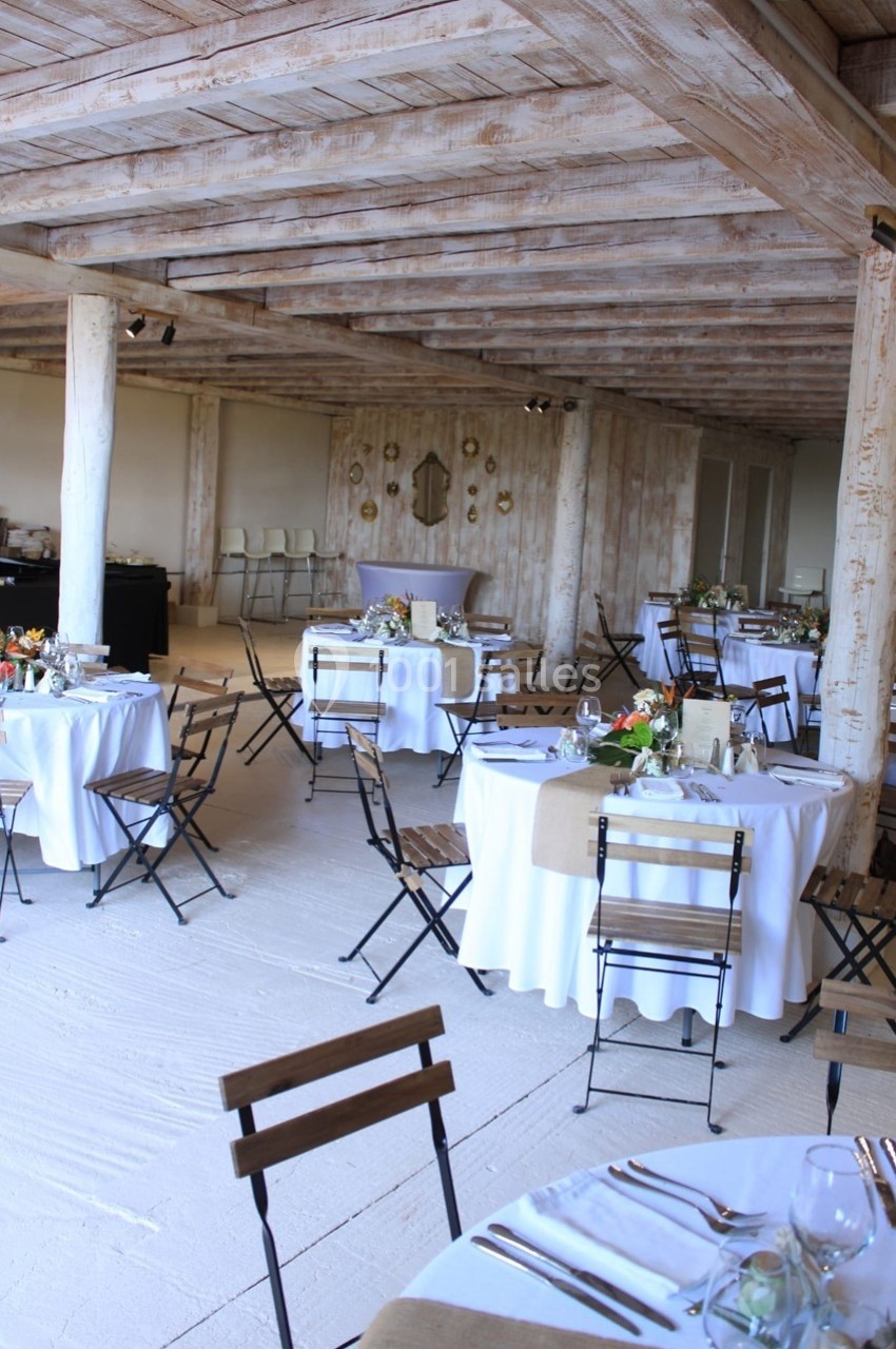 Salle de réception lumineuse avec tables rondes décorées, chaises pliantes et poutres en bois apparentes.