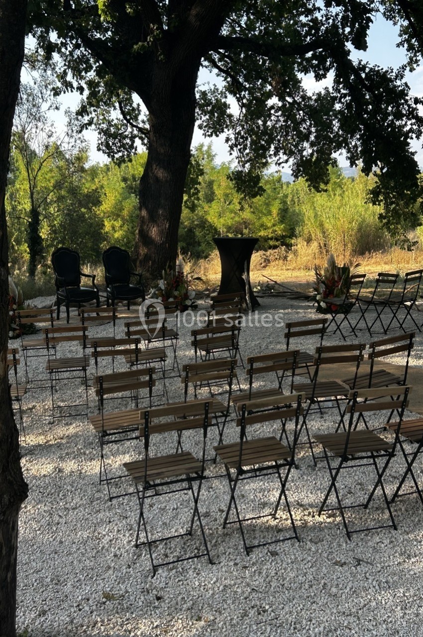 Chaises pliantes disposées en rangées sous un grand arbre, formant un espace extérieur pour une cérémonie.