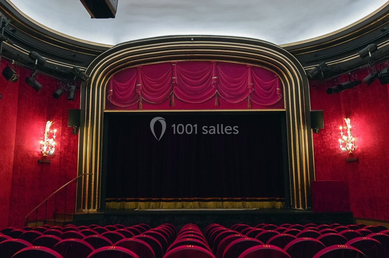 Salle de théâtre avec rideaux rouges, scène vide et rangées de sièges en velours rouge.