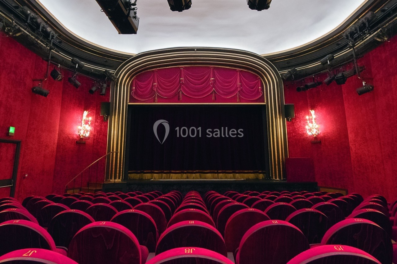 Salle de théâtre vide avec des sièges rouges, une scène encadrée de rideaux et un éclairage tamisé.