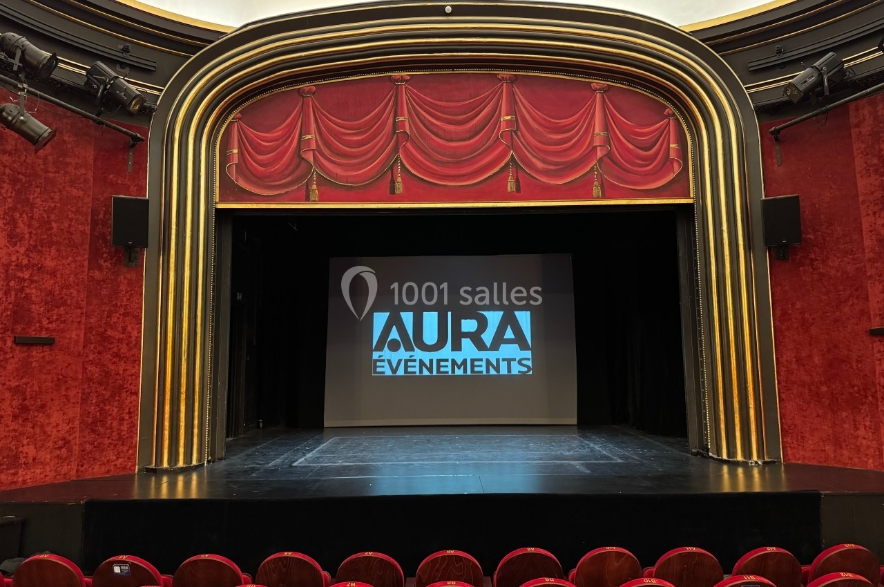 Scène de théâtre avec rideaux rouges, écran central affichant ’Aura Événements’, et rangées de sièges vides au premier plan.