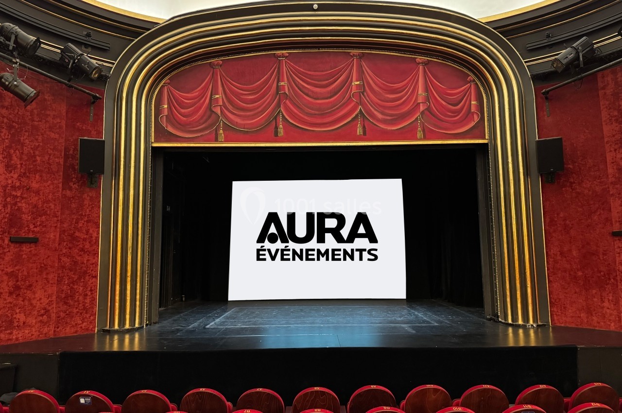 Scène de théâtre avec rideaux rouges, écran affichant ’Aura Événements’ et rangées de sièges vides au premier plan.