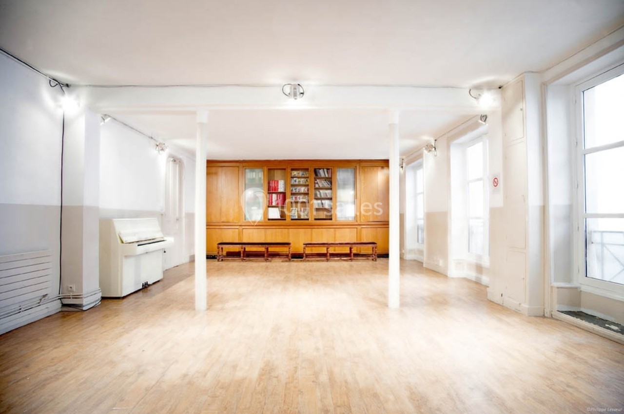 Salle lumineuse avec parquet clair, murs blancs, bibliothèque en bois, piano et grandes fenêtres laissant entrer la lumière.