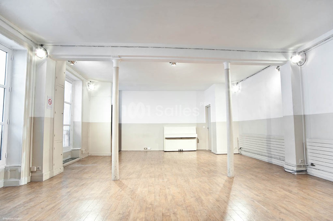 Salle vide avec parquet en bois clair, murs blancs et beiges, colonnes centrales et grandes fenêtres laissant entrer la…