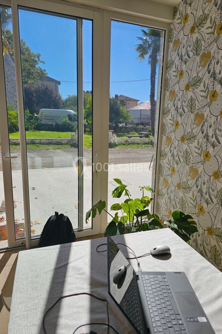 Vue depuis une pièce lumineuse avec une table, un ordinateur portable et une baie vitrée donnant sur un jardin ensoleillé.