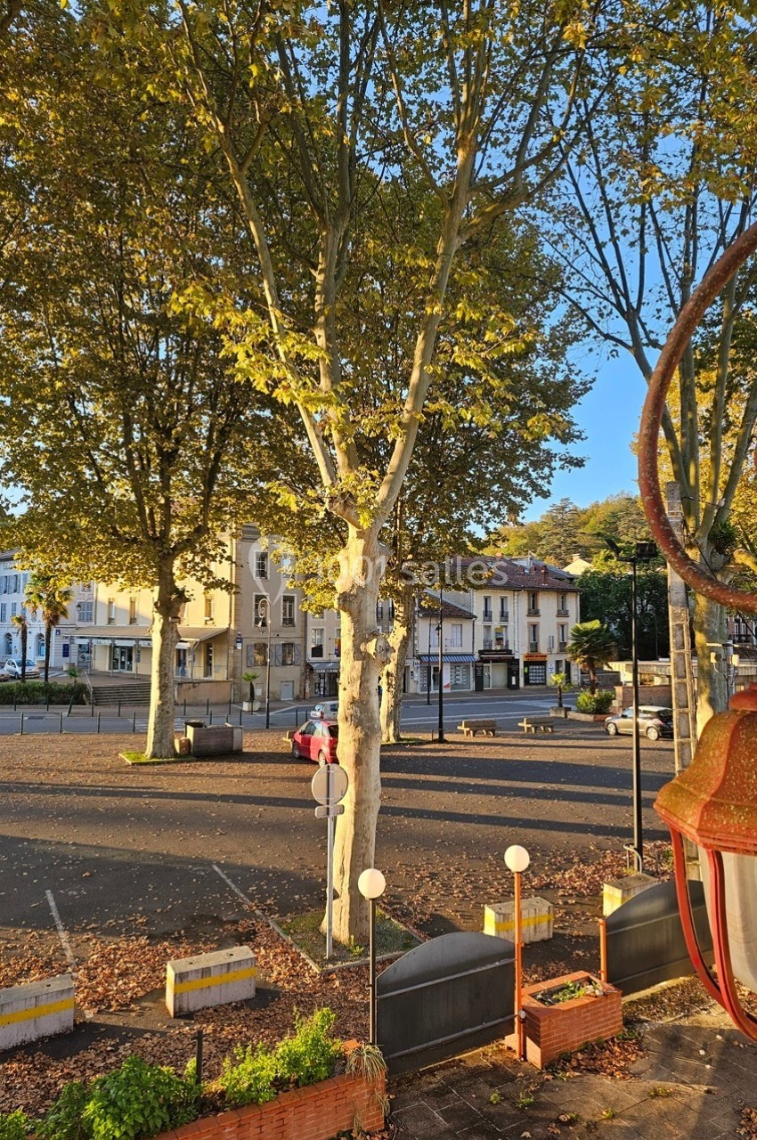 Place bordée d'arbres et de bâtiments sous une lumière dorée, avec des lampadaires et des jardinières au premier plan.