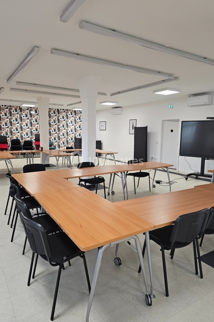 Salle de réunion lumineuse avec tables en bois disposées en U, chaises noires et écran de présentation.