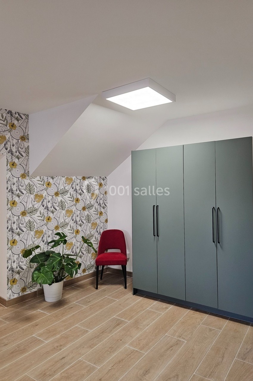 Chambre lumineuse avec armoire grise, chaise rouge, plante en pot et mur décoré de papier peint floral.