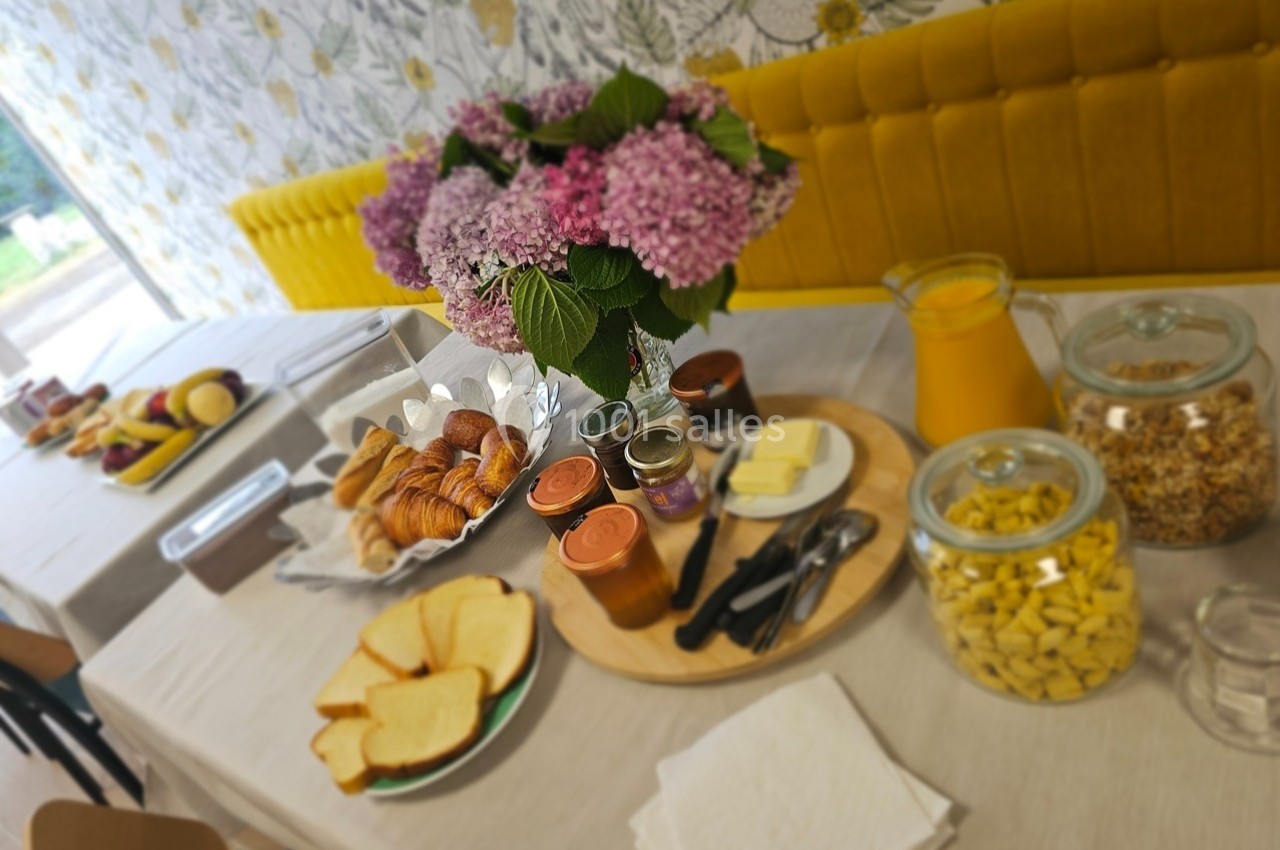 Petit-déjeuner servi sur une table avec viennoiseries, pain, confitures, céréales, fruits, jus d'orange et bouquet de fleurs.