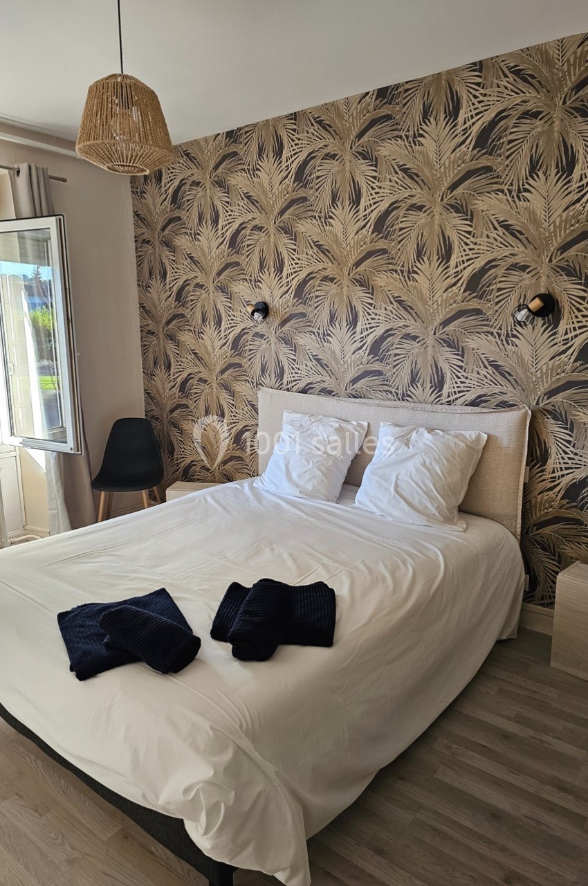 Lit double avec draps blancs, deux serviettes pliées, mur à motif végétal, fenêtre ouverte et chaise noire.