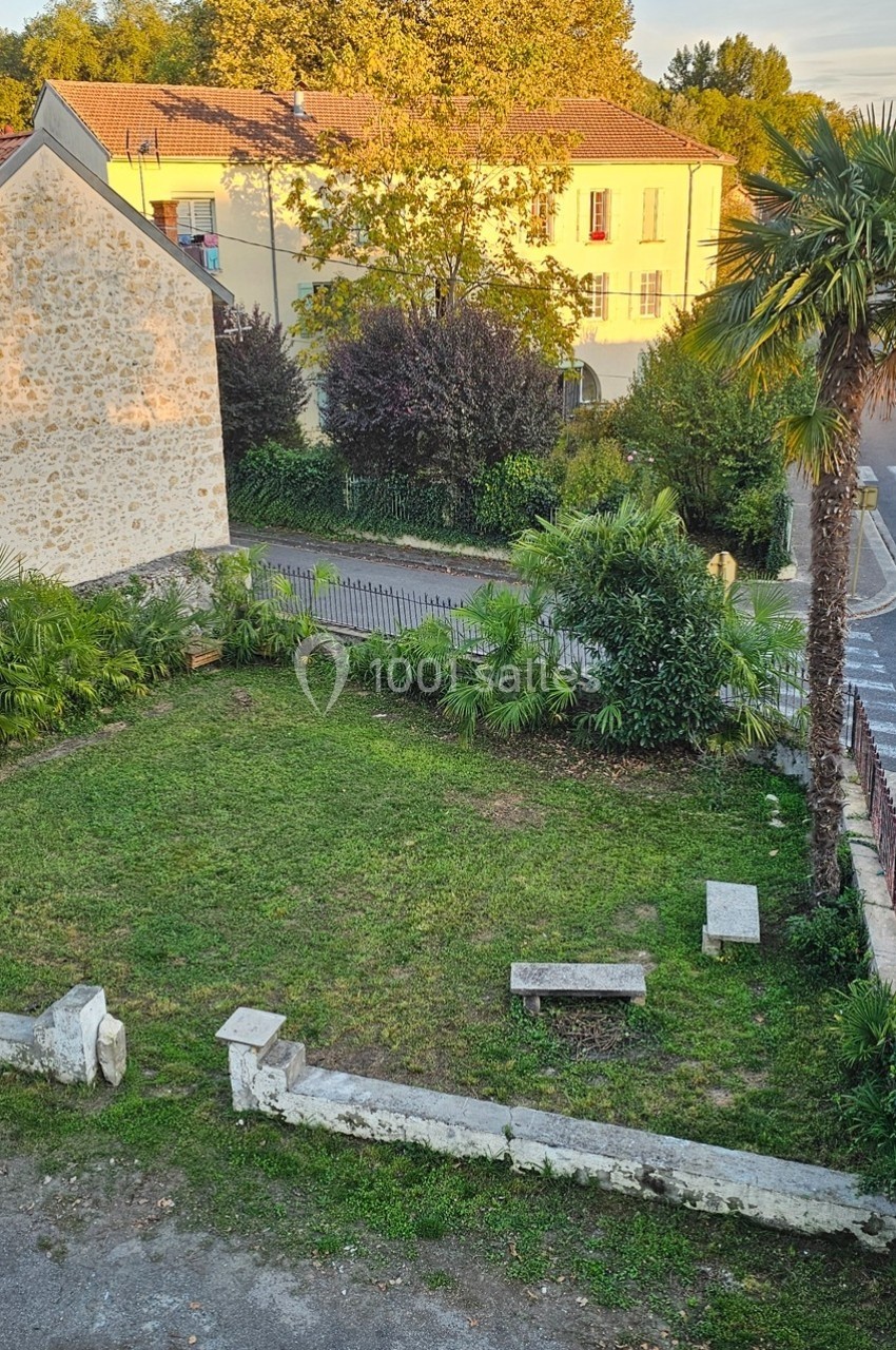 Petit jardin avec pelouse, bancs en pierre, palmiers et arbres, entouré de bâtiments en pierre et végétation.