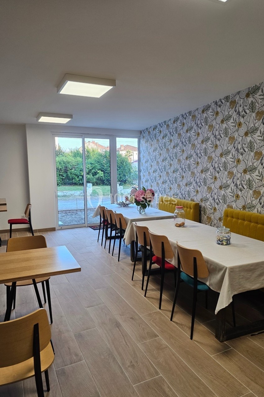 Salle lumineuse avec tables en bois, chaises colorées, banquette jaune et décoration murale à motifs floraux.