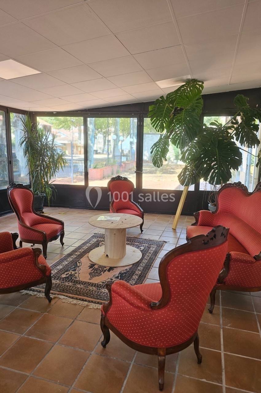 Salon avec fauteuils et canapé rouges autour d'une table basse, plantes vertes et grandes fenêtres donnant sur l'extérieur.