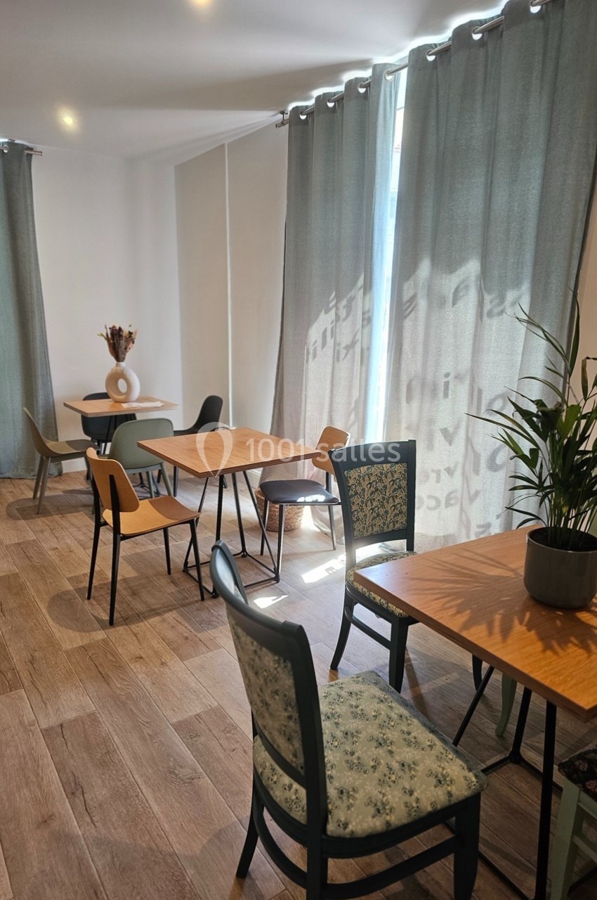 Salle lumineuse avec tables en bois, chaises variées et rideaux gris laissant passer la lumière naturelle.