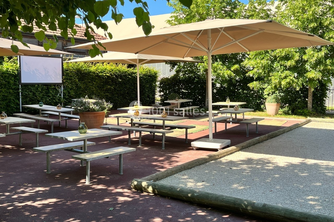 Terrasse extérieure avec tables, bancs, parasols et écran, entourée de verdure et baignée de lumière naturelle.