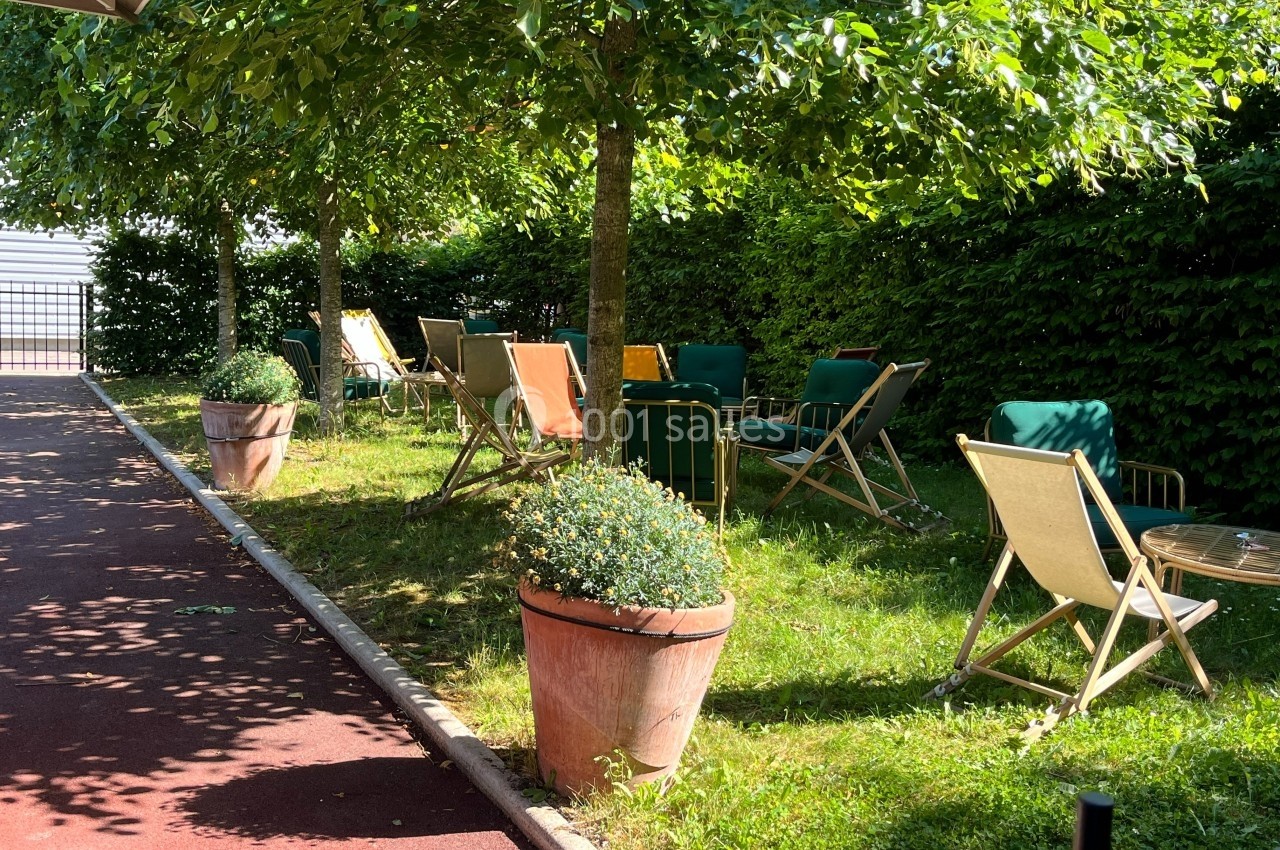 Chaises longues colorées disposées sous des arbres dans un espace ombragé, entouré de verdure et de pots de fleurs.