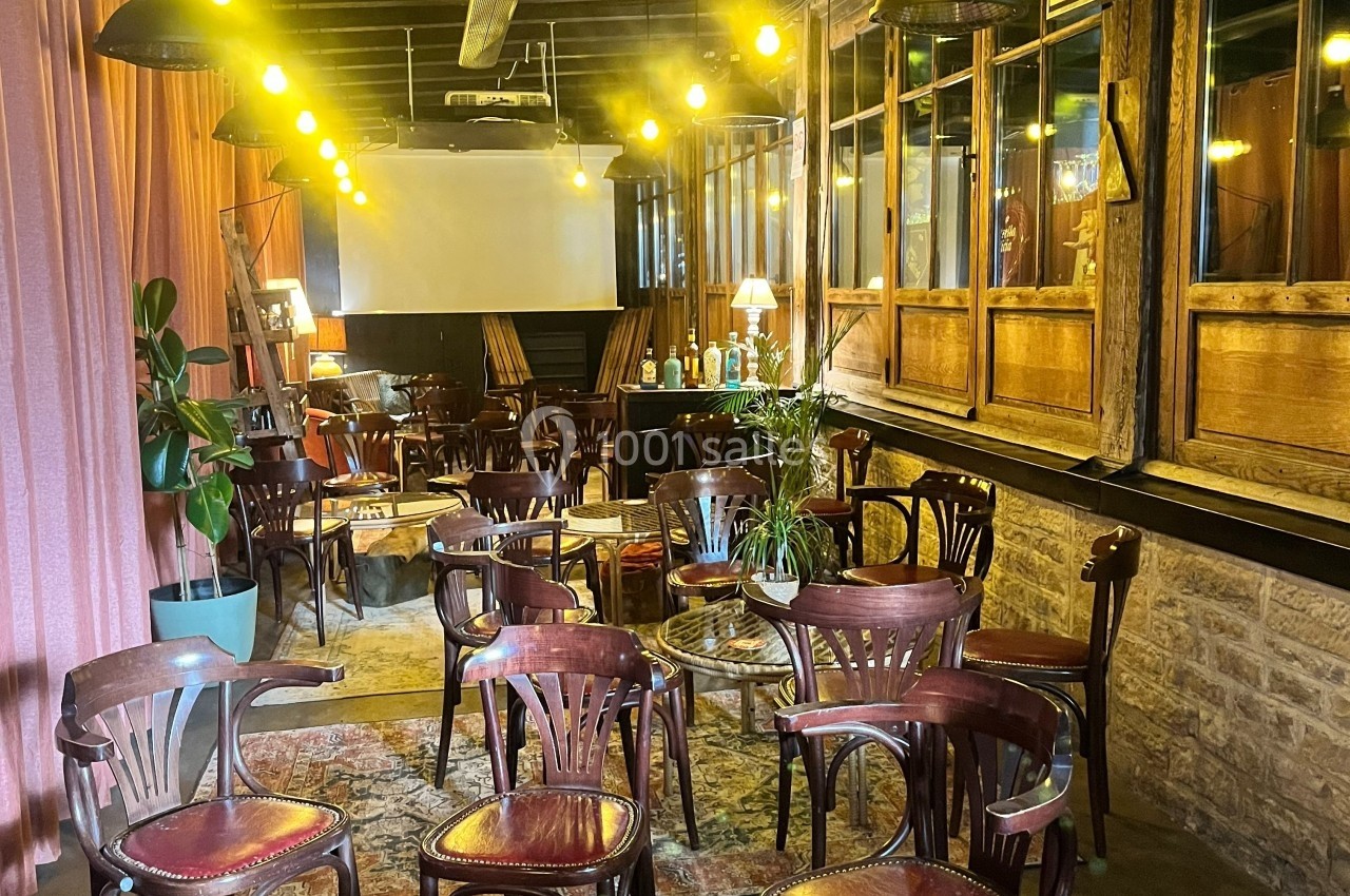 Salle aménagée avec des chaises en bois, des tables basses, des plantes et un écran de projection au fond.