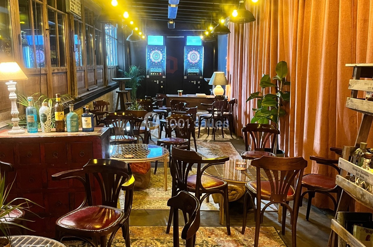 Salle chaleureuse avec des tables et chaises en bois, éclairage tamisé, plantes et jeux de fléchettes au fond.