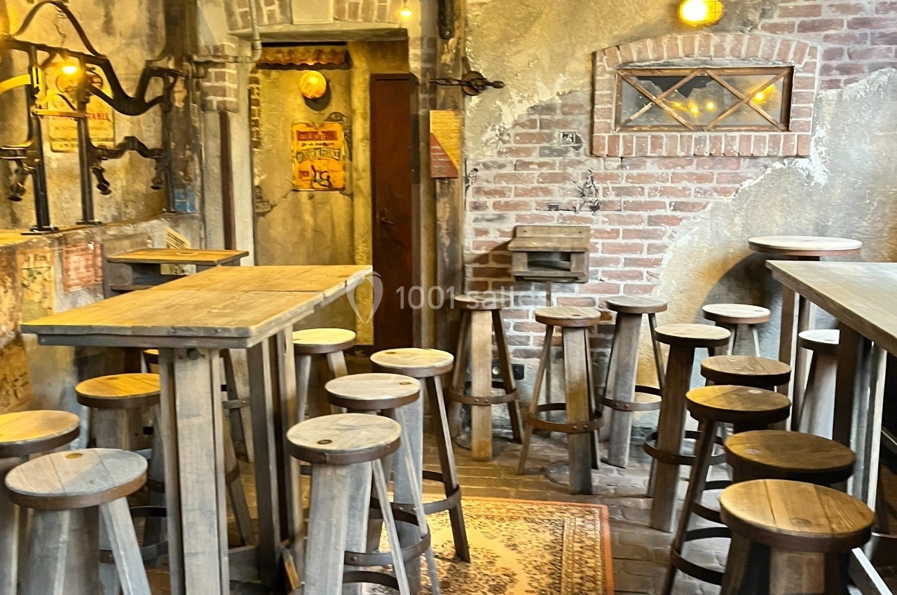 Salle rustique avec murs en briques, tabourets en bois autour de tables hautes et éclairage chaleureux.