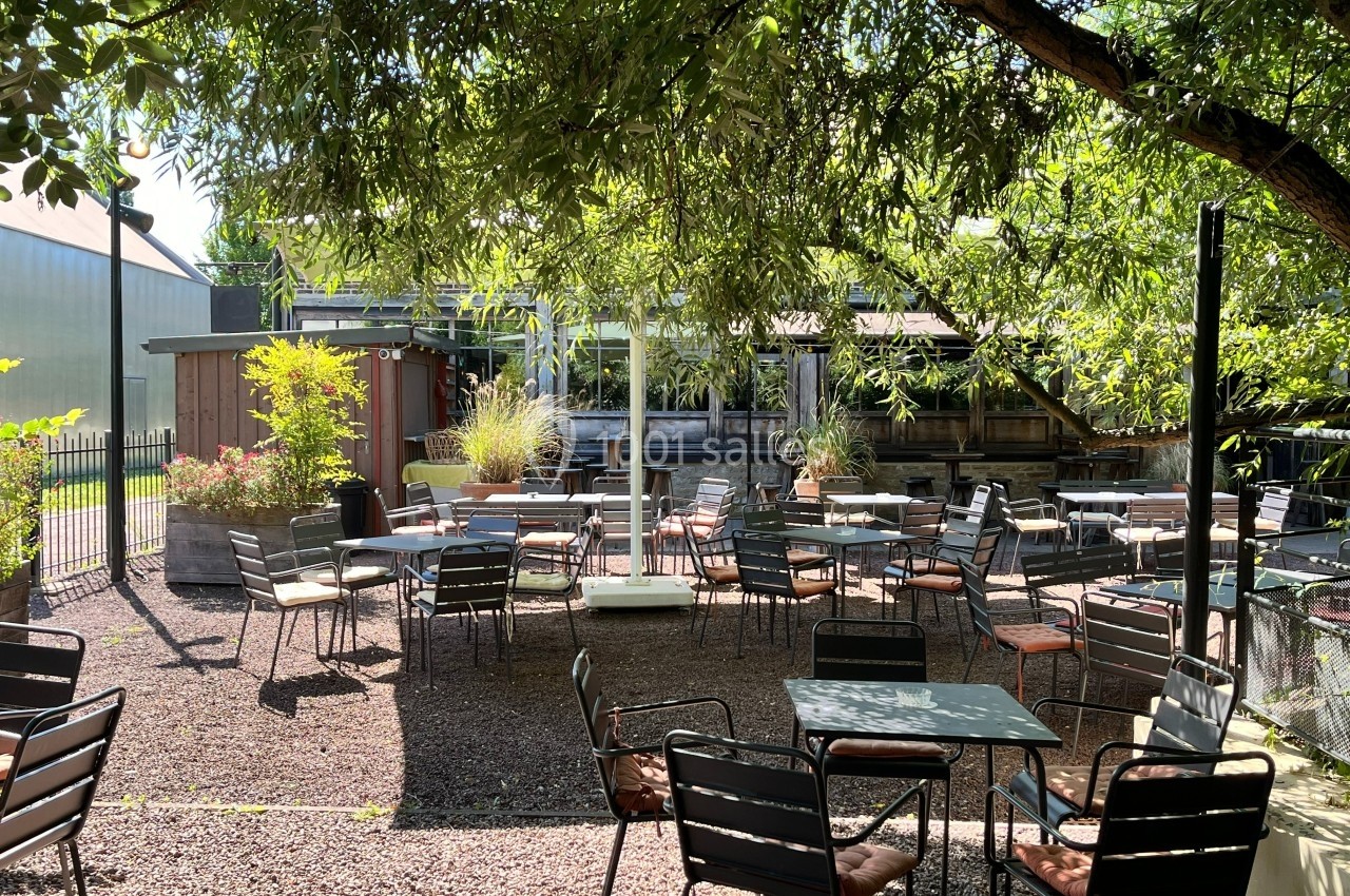 Terrasse extérieure ombragée avec des tables et des chaises, entourée de végétation et baignée de lumière naturelle.