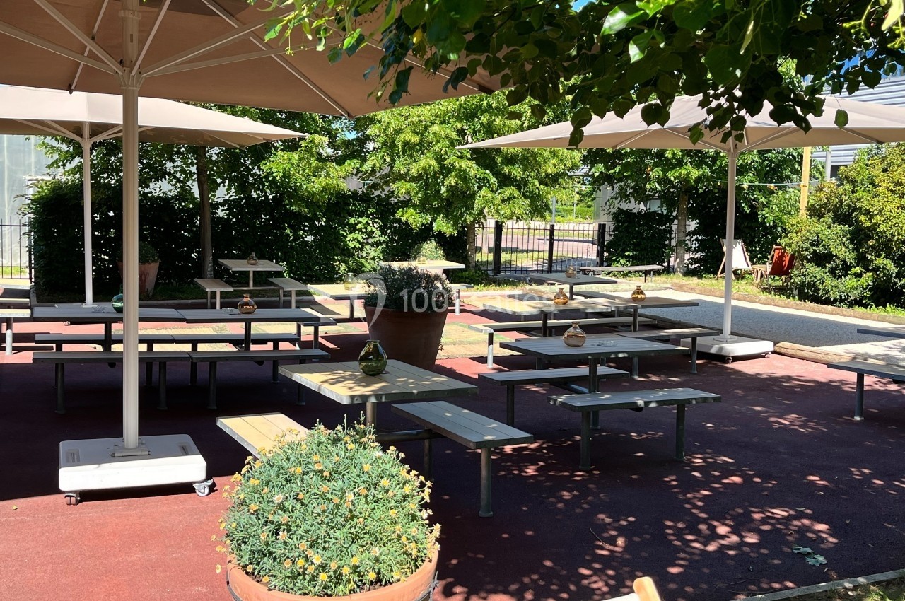 Terrasse extérieure avec tables et bancs sous des parasols, entourée de verdure et de plantes en pots.