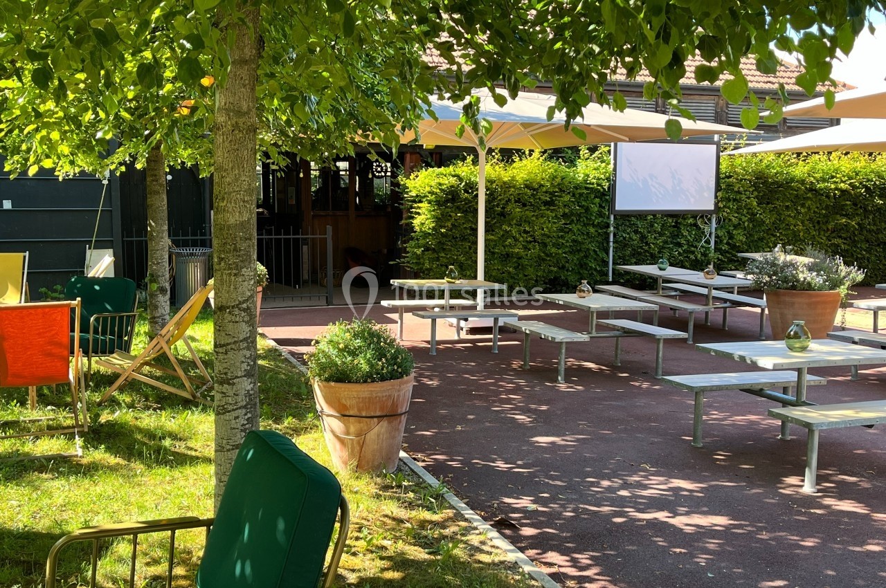 Terrasse extérieure ombragée avec tables, chaises et parasols, entourée de verdure et de plantes en pots.