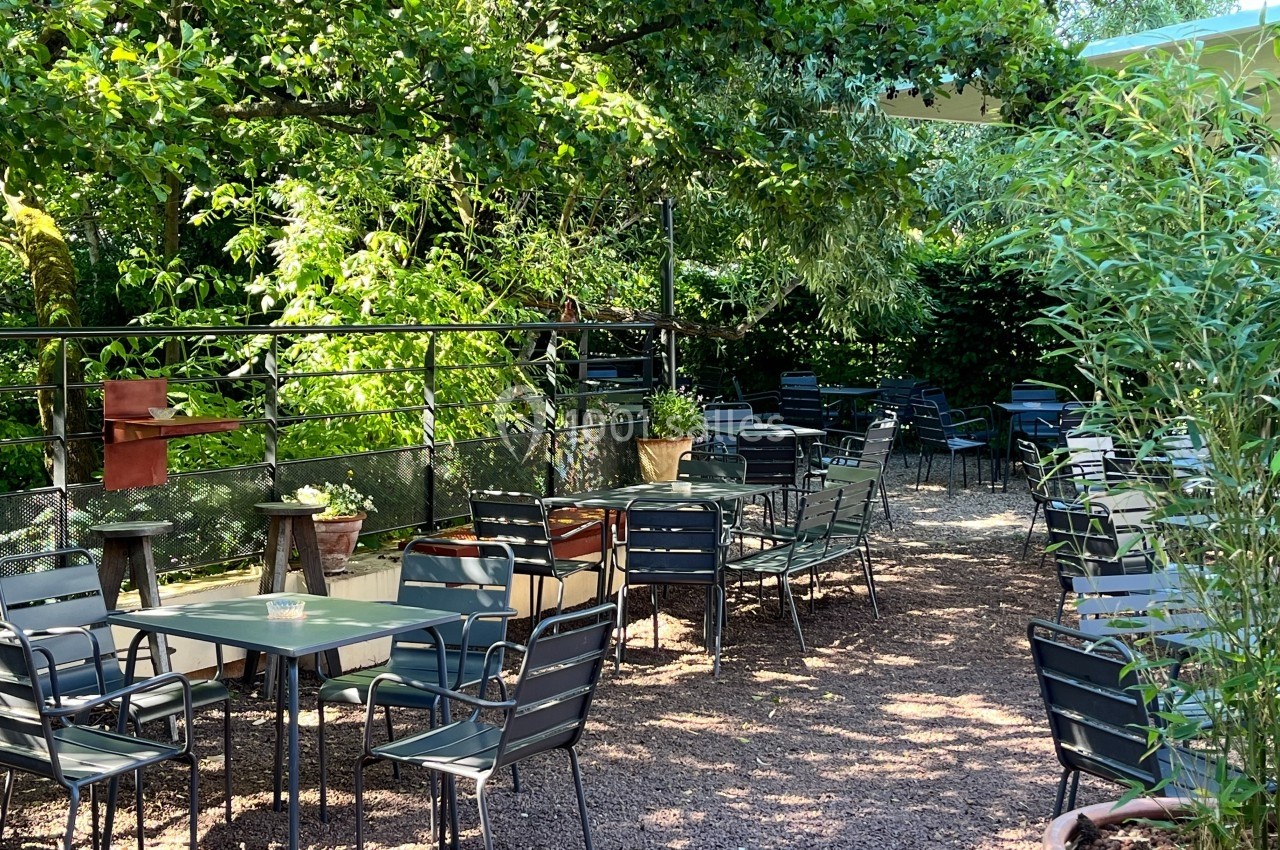 Terrasse extérieure avec tables et chaises en métal, entourée de végétation dense et ombragée.