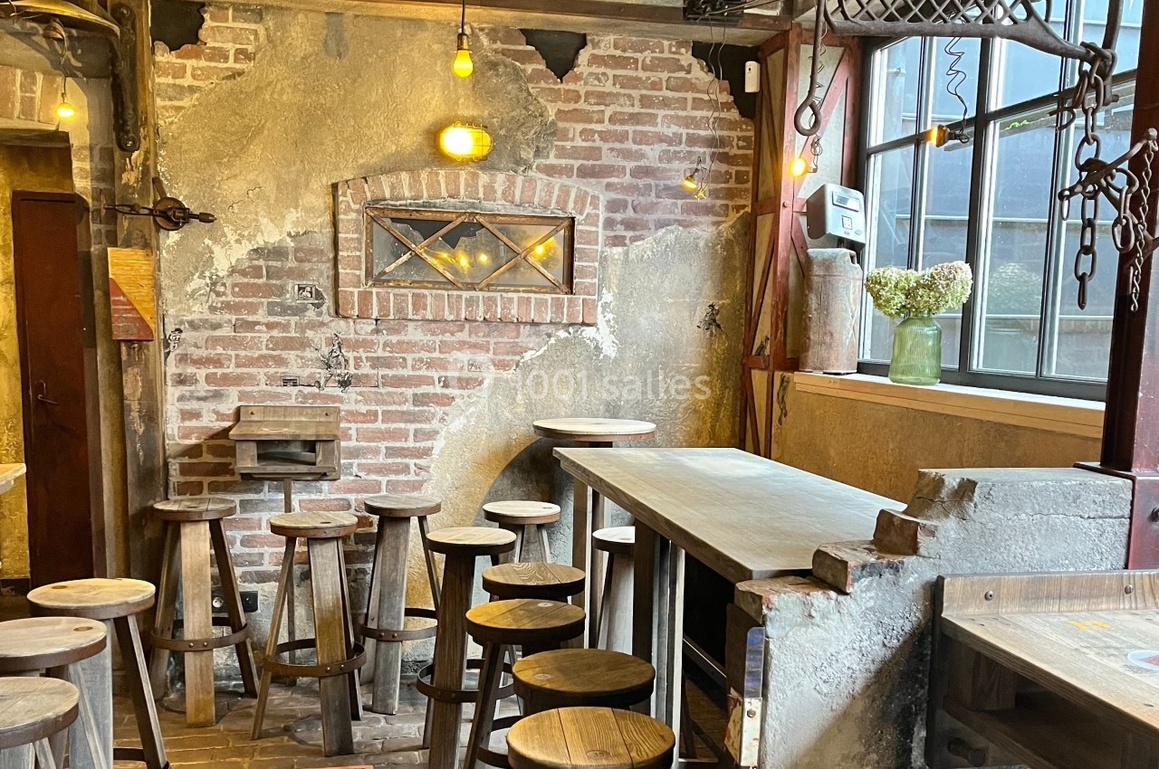 Intérieur d'un café rustique avec murs en briques, tabourets en bois, table haute et fenêtre laissant entrer la lumière.