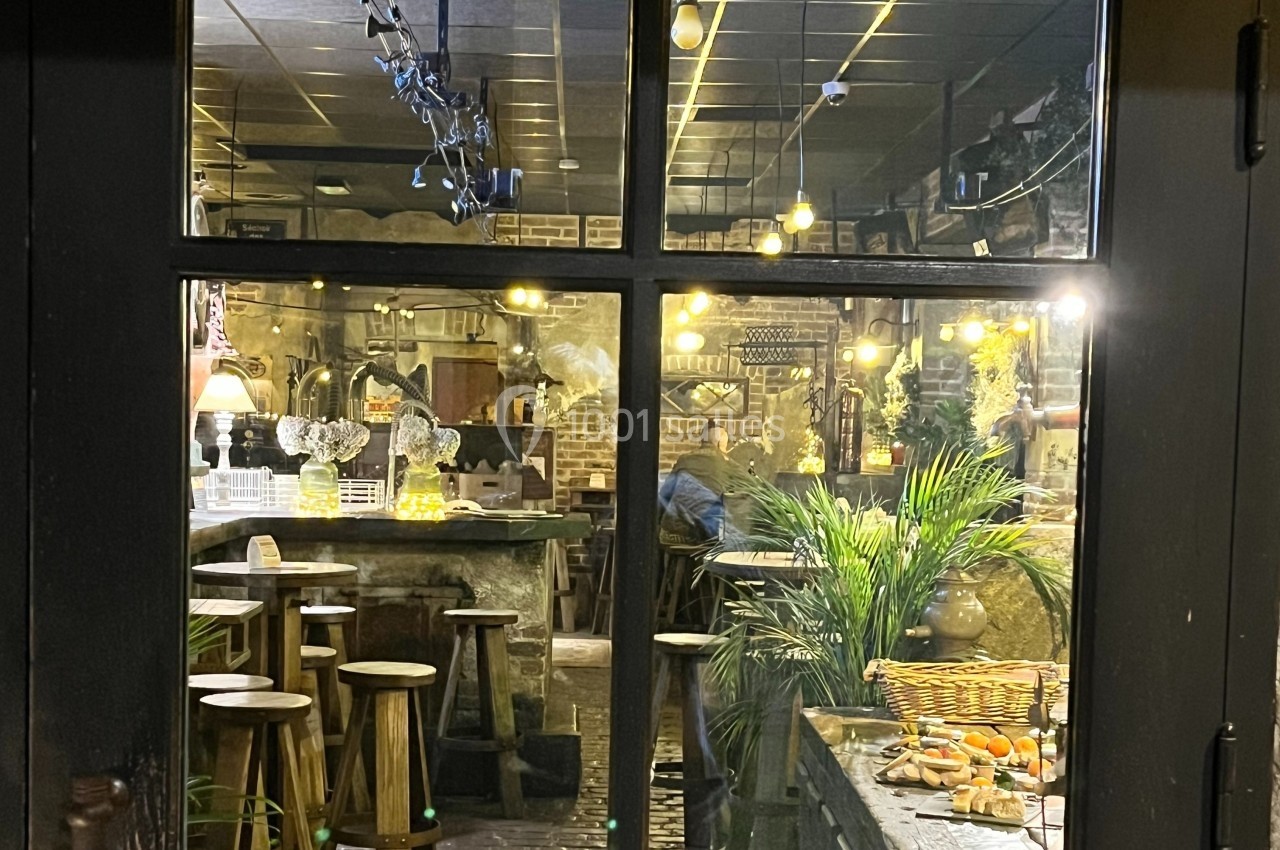 Vue à travers une vitre d'un intérieur de restaurant chaleureux avec tabourets en bois, plantes et éclairage tamisé.