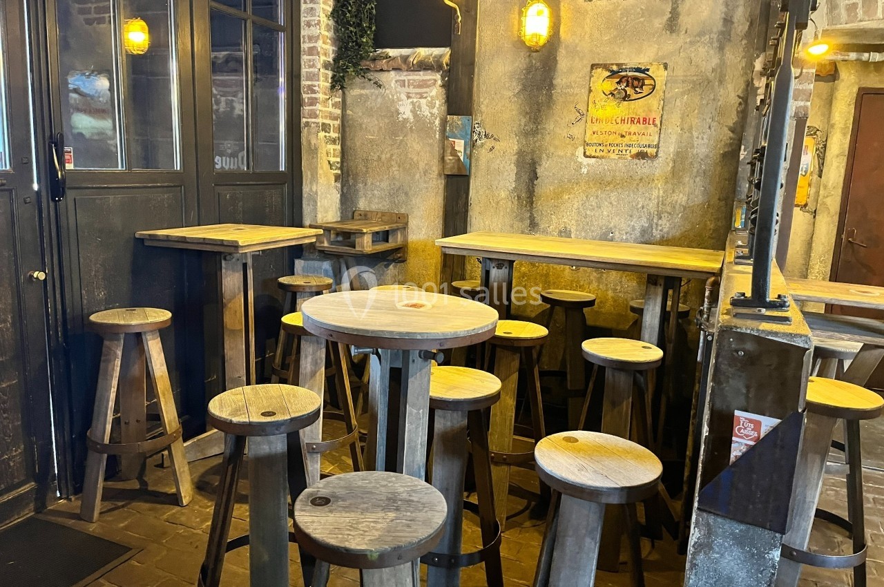 Intérieur d'un bar rustique avec tabourets en bois, tables hautes et murs en pierre décorés d'affiches vintage.