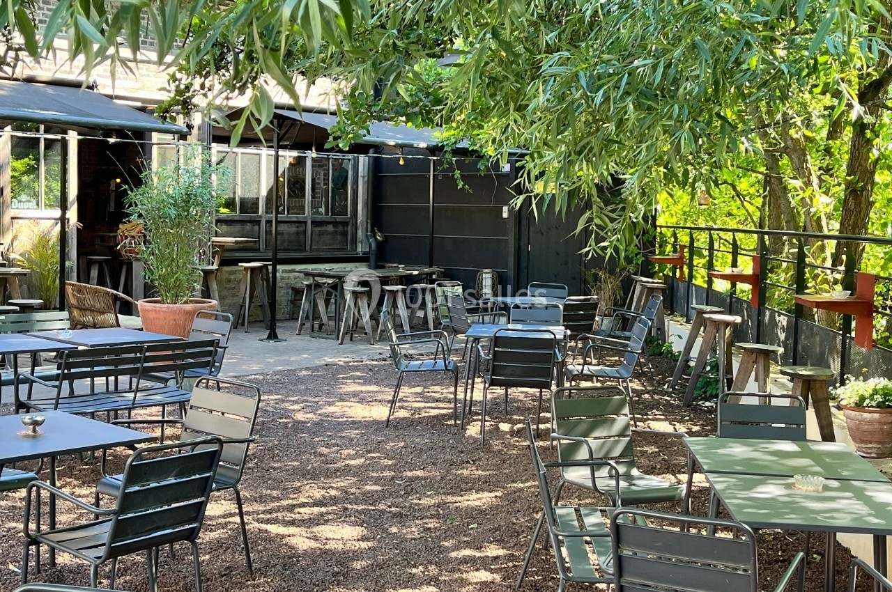 Terrasse extérieure ombragée avec tables et chaises en métal, entourée de végétation et éclairée par la lumière naturelle.