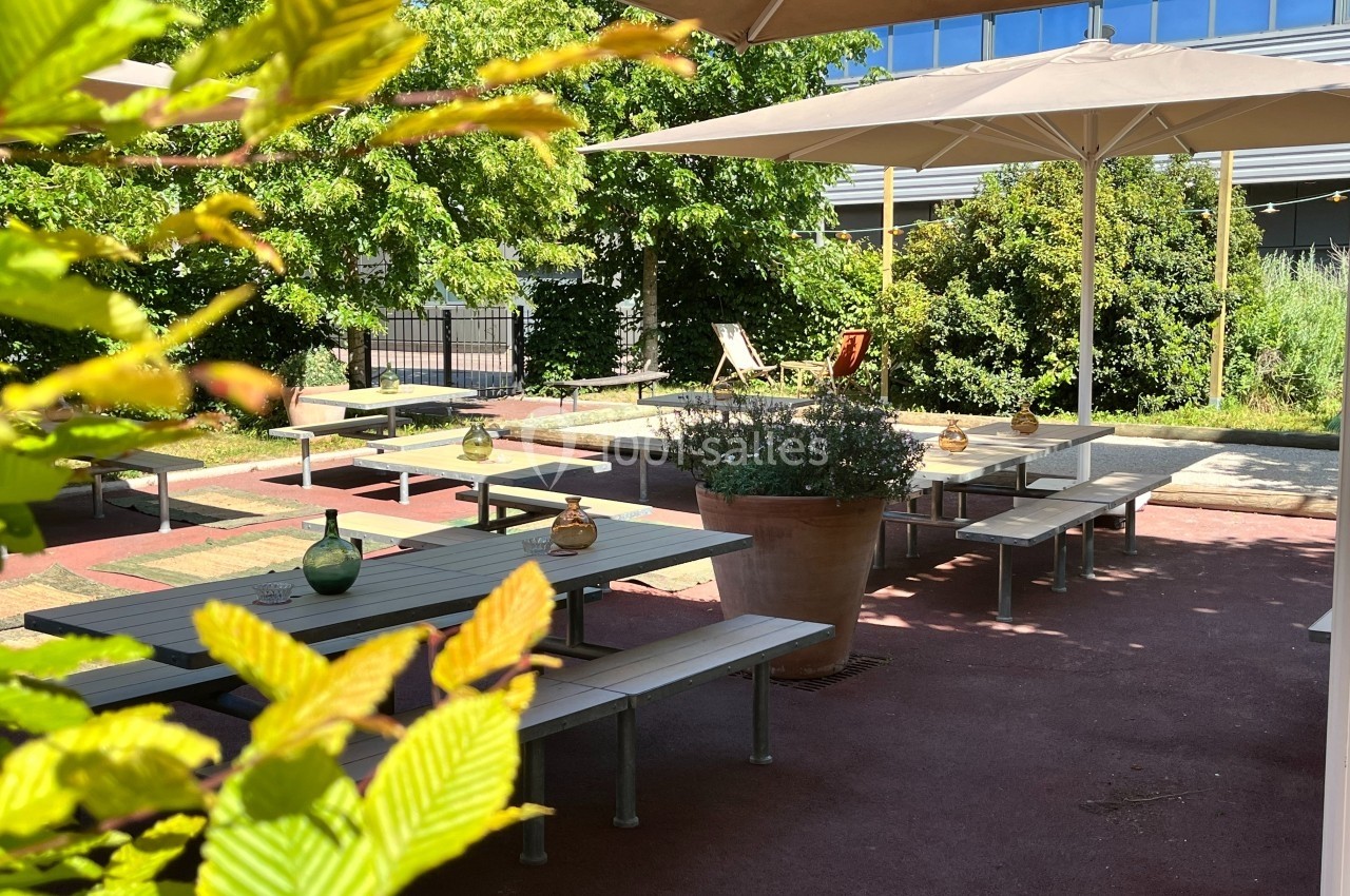 Terrasse extérieure avec tables, bancs, parasols et végétation environnante sous un ciel ensoleillé.