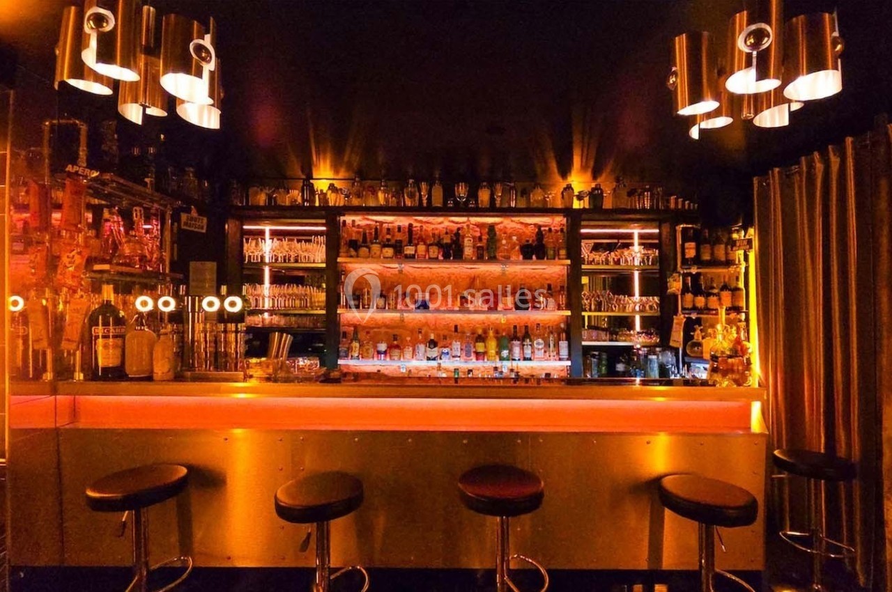 Bar avec comptoir éclairé, tabourets noirs, étagères remplies de bouteilles et ambiance lumineuse tamisée.