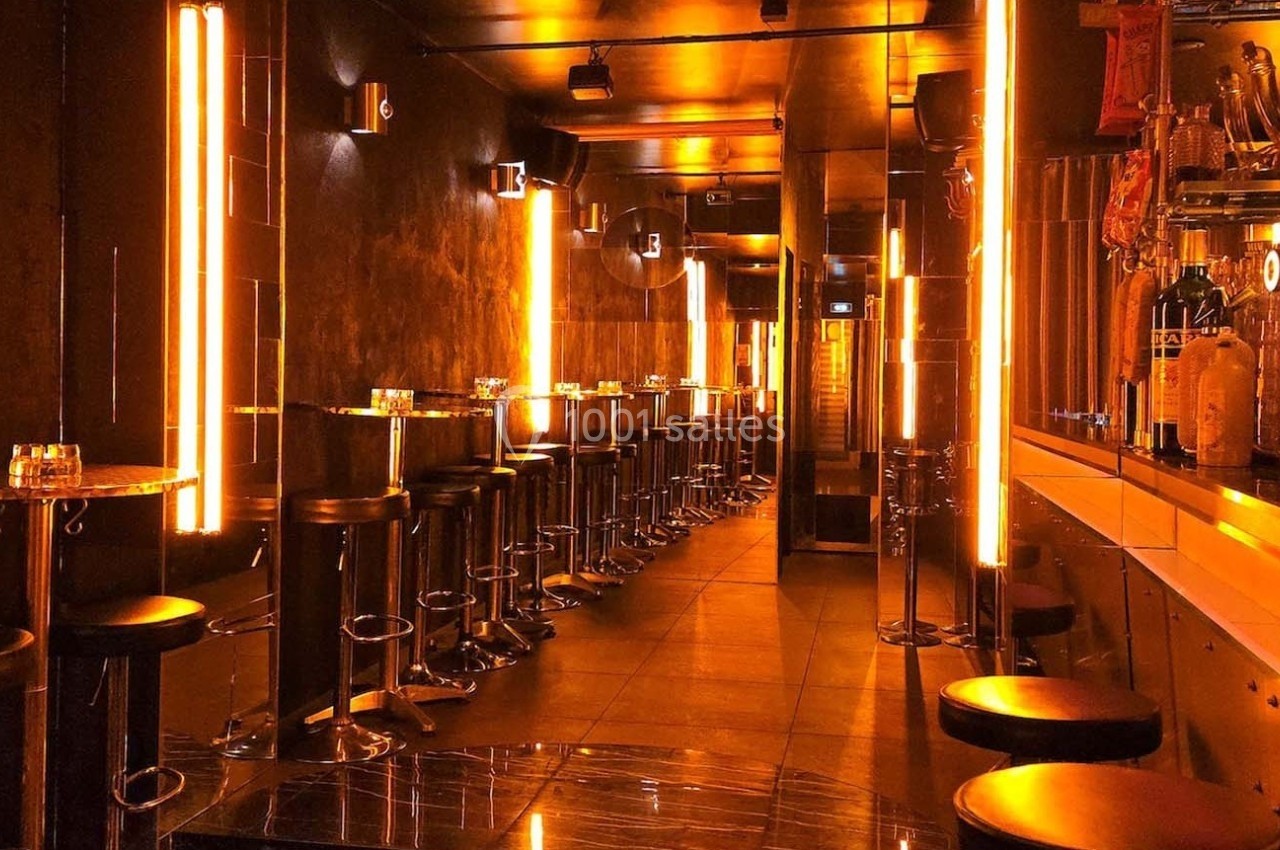 Intérieur d'un bar moderne avec éclairage orange, tabourets hauts alignés et comptoir sur la droite.
